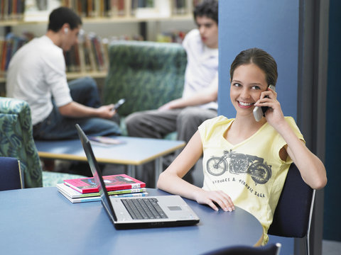 Happy teenage girl using cellphone with laptop on table in library
