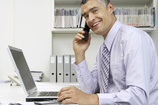 Portrait of smiling middle aged businessman with laptop using cellphone at desk