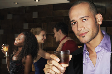 Portrait of a young man holding drink with people dancing behind in the bar