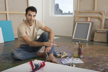 Portrait of a male artist sitting with painting tools on floor at a studio