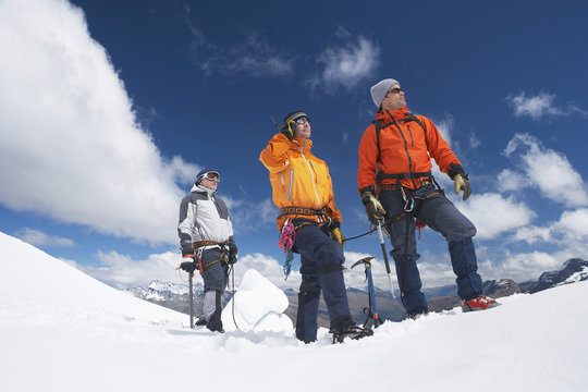 Three male mountain climbers reaching snowy peak against clouds