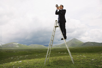 Full length of businessman on ladder speaking through megaphone in mountain field