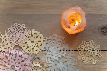 Christmas still life. Candlestick of pink salt with burning candle and a lot of snowflakes cut out of paper on light wooden surface. Close up, top view.
