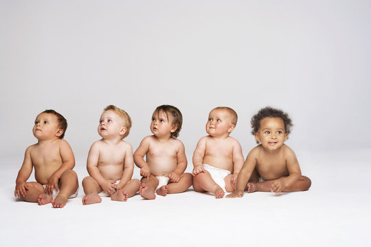 Row of multiethnic babies sitting side by side looking away isolated on gray background