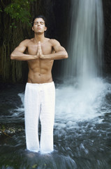 Young man with hands clasped meditating while standing against waterfall