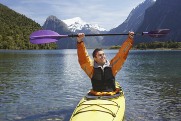 Cheerful man holding up kayak oar over head in mountain lake
