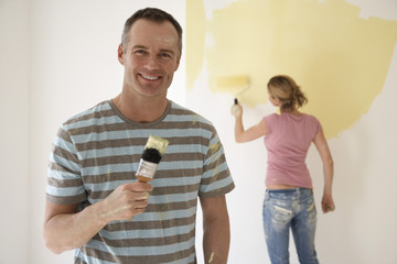 Smiling man holding paintbrush while woman paints wall with roller in the background