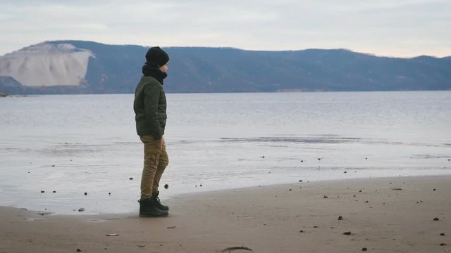 Boy dressed warmly strolls along the beach. It warm jacket, hat, scarf, pants and boots Lightstone color. Against the background of a view. Beautiful sea, captivating mountains, low wave.