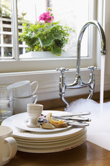 Closeup of a stack of dirty dishes and silverware by sink with running water below window with...