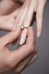 Closeup of man placing engagement ring in woman's finger against gray background