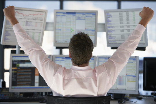 Rear view of stock trader with hands raised looking at multiple computer screens