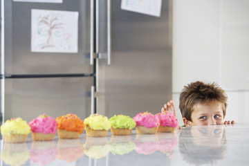 Happy young boy peaking over counter at row of cupcakes in kitchen