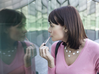 Young woman applying lip gloss while looking at reflection of self in window