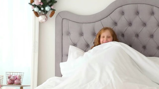 Portrait of cute funny little girl on Christmas morning in white home interior. Child pretending to be sleeping than rises cheerfully in bed. Real time full hd video footage.