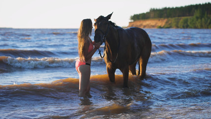 A beautiful blonde young sexy woman standing next to a horse at a lake