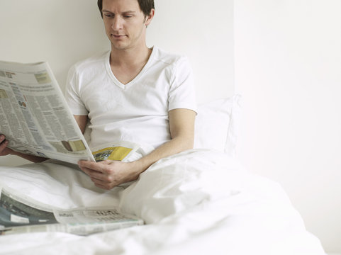 Young Man Reading Newspaper In Bed