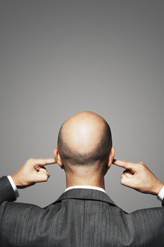Rear view of a bald businessman with fingers in ears against gray background