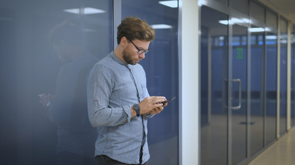 Serious man standing at the wall in an office and writing sms on mobile phone.