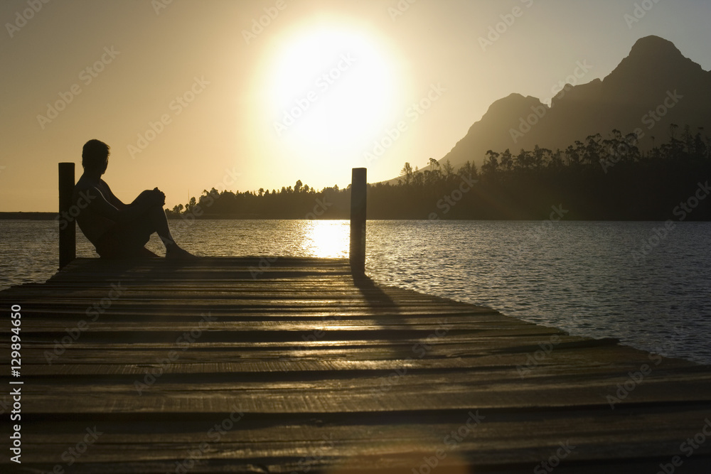 Wall mural side view of man sitting on dock by lake enjoying sunset