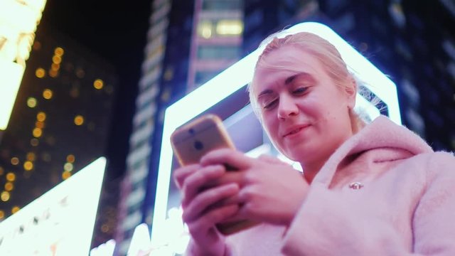 Times Square, New York. Portrait of a young caucasian woman on the background of skyscrapers at night. Smiling enjoys smartphone
