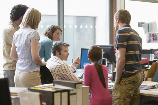Rear view of a group of multiethnic office workers around colleague using computer