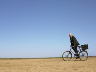 Side view of middle aged businessman with briefcase riding bicycle in desert