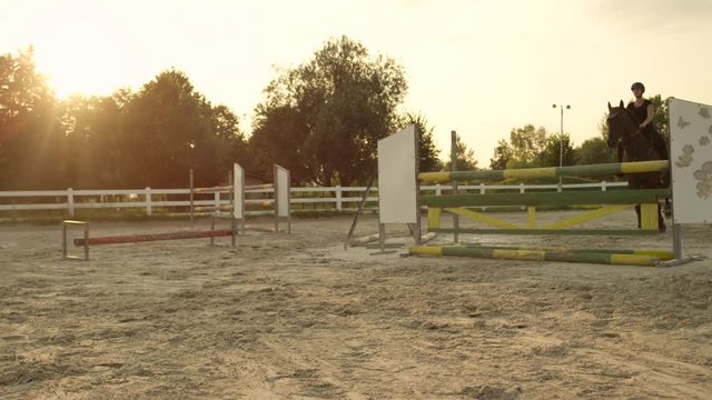CLOSE UP: Young woman training horse jumping on outdoors arena at sunset