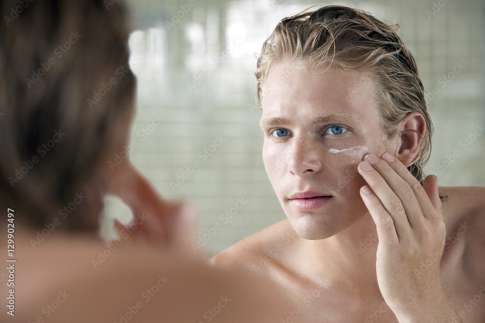 Wall mural Closeup of handsome young man applying facial cream in front of mirror