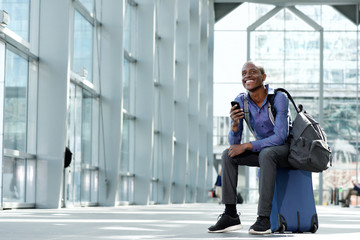 smiling businessman sitting on luggage at airport with cell phone