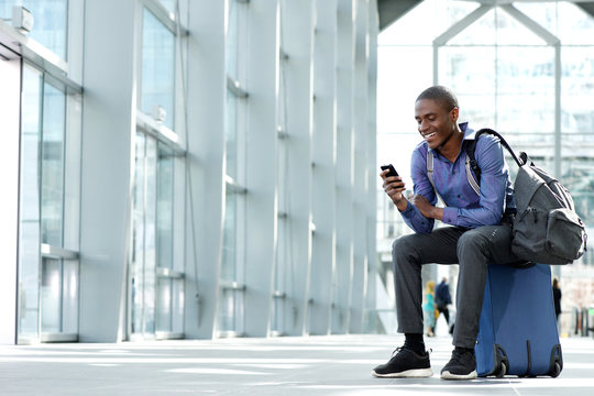 Smiling Young Businessman Sitting On Suitcase Looking At Phone