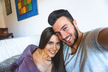Fototapeta na wymiar Stunning happy couple taking a selfie while relaxing at home - Boyfriend and girlfriend taking a selfie on a white sofa in their apartment