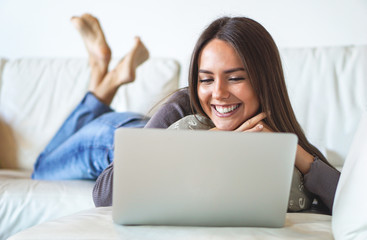 Naklejka premium Beautiful smiling woman laying on a sofa working on her laptop - Pretty girl watching something interesting on her computer - Stunning girl talking to her boyfriend on webcam