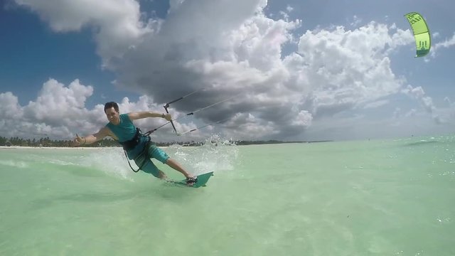 SLOW MOTION: Smiling kiter kiteboarding past the camera showing shaka surf sign