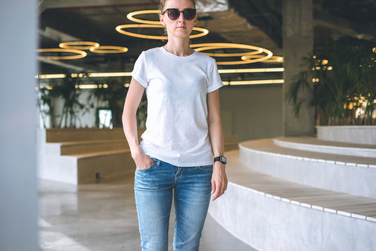 Front view.Young woman in sunglasses,white t-shirt and blue jeans standing in room with modern interior, with his hand in pocket of jeans. Mock up.In background, seating and lighting in form of rings.