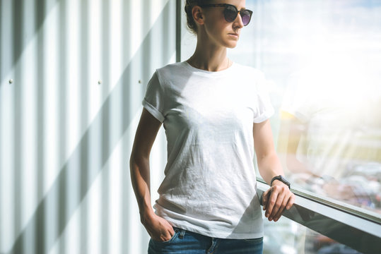 Front view. Young woman in sunglasses,white t-shirt and jeans standing near window in room, with his hand in pocket of jeans. In background white wall. Girl looks out window.Mock up.