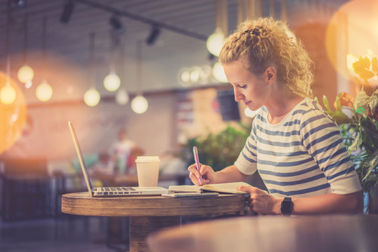 Side view. Young woman in striped blouse sitting at table in coffee shop and makes notes in notebook. In front of her is laptop next to cup of coffee. Online learning. Student doing homework.