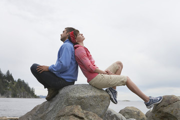 Full length side view of a young couple sitting back to back on rocks against ocean