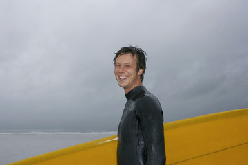 Side view of cheerful young man with surfboard against cloudy sky at beach