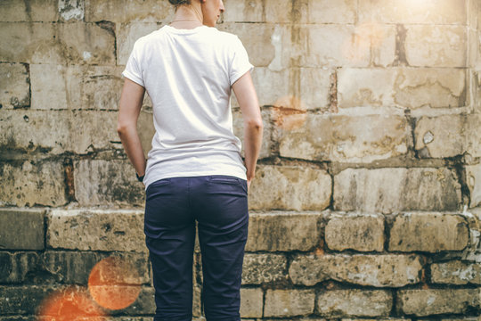 Summer day. Back view. Young woman in a white T-shirt and blue pants standing on a background of a stone wall with his hands in his pockets. Cropped image. Mock up.