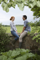 Full length side view of a smiling couple sitting on countryside wall against landscape