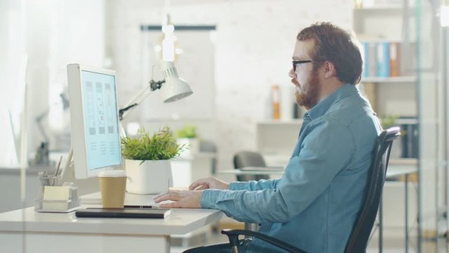 Young Bearded Man Working on Personal Computer while Sitting in His Creative Agency Office. Shot on RED Cinema Camera in 4K (UHD).