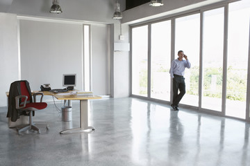 Full length of a male executive using cellphone against glass wall in empty office