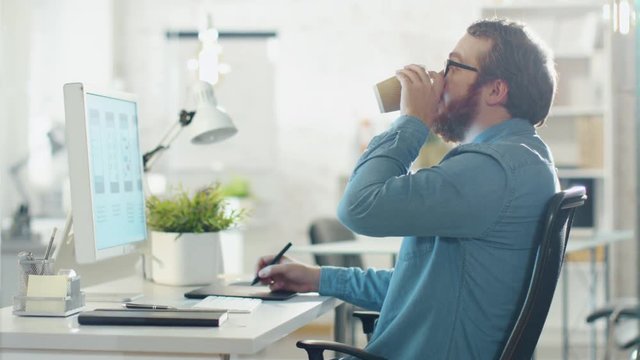 Young Bearded Man Taking Notes while Working on Desktop Computer. He Sips Coffee. He Sits in a Creatively Designed Office. Shot on RED Cinema Camera in 4K (UHD).
