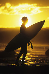 Side view of a silhouette male surfer carrying surfboard along beach at sunset