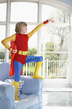 Full length of a young boy in superman costume standing on armchair and pointing up