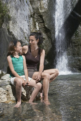 Smiling mother and daughter sitting on rock by waterfall