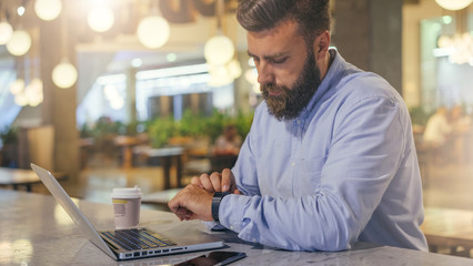 Side view.Young bearded businessman wearing blue shirt, sitting at table in cafe and uses smartwatch. Nearby is laptop, smartphone and cup of coffee. Man checks messages on digital gadget.Film effect.