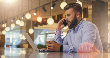 Side view.Young bearded businessman wearing blue shirt, sitting at table in cafe,talking on cell phone while holding cup of coffee and looking at laptop screen. Man works outside office, using gadget.