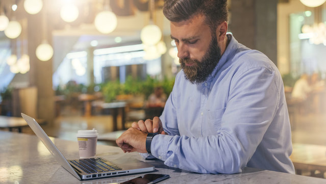 Side view.Young bearded businessman wearing blue shirt, sitting at table in cafe and uses smartwatch. Nearby is laptop, smartphone and cup of coffee. Man checks messages on digital gadget.Film effect.