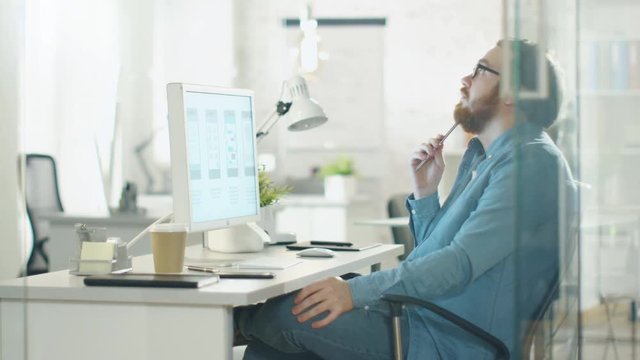 Focused Creative Man Thinks while Sitting at His Workplace Desk. He Holds Pencil and Looks at Desktop Computer Screen. Shot on RED Cinema Camera in 4K (UHD).